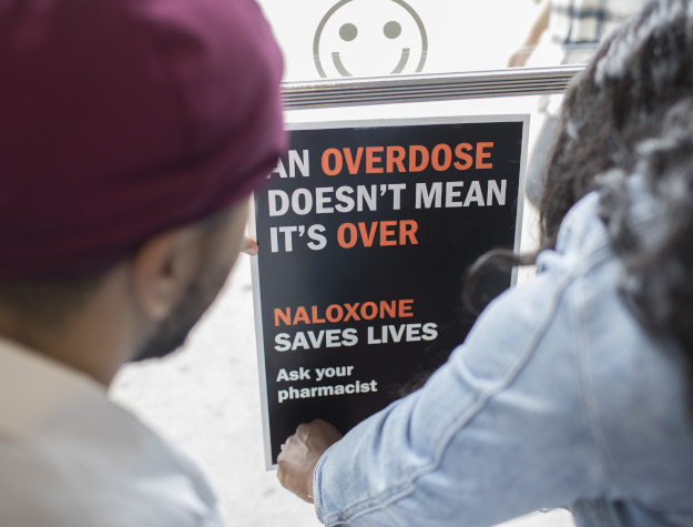 Two people observing a sign with text 'An overdose doesn't mean it's over. Naloxone saves lives. Ask your pharmacist