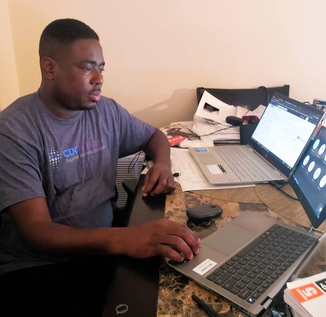 A black man seated at a desk and wearing a CDC Foundation tshirt focuses on three computer screens in front of him.