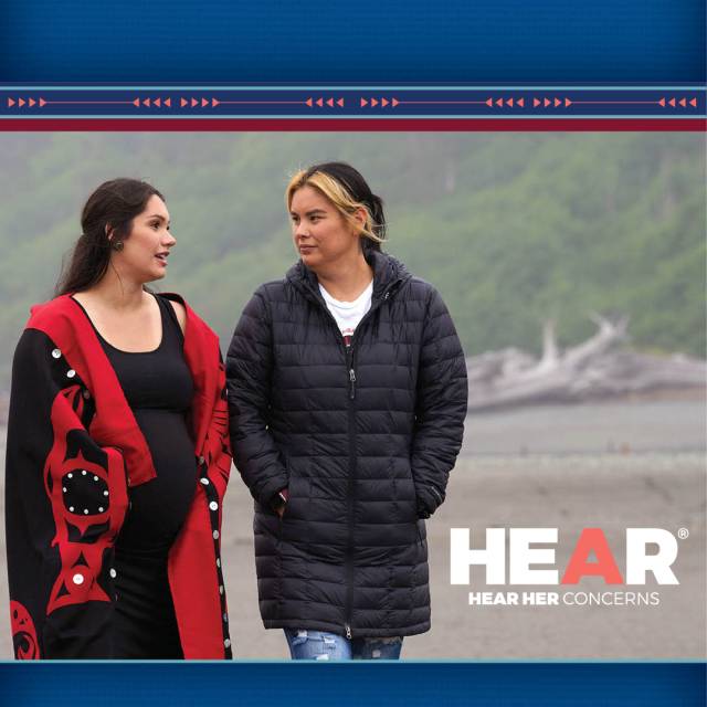 A pregnant woman in a red sweater walks on the beach with a friend wearing an overcoast