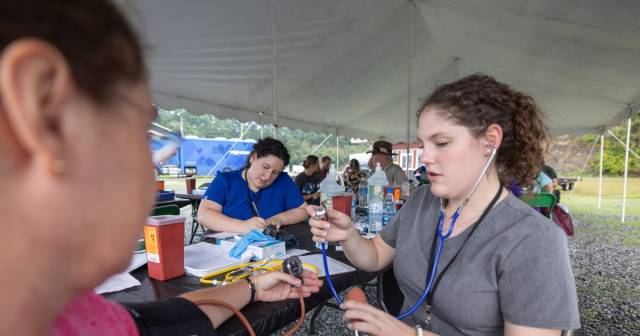 A woman with a stethoscope takes a person's blood pressure under a tent.