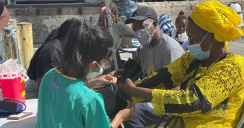 A nurse in green scrubs puts a blood pressure cuff on a woman in yellow