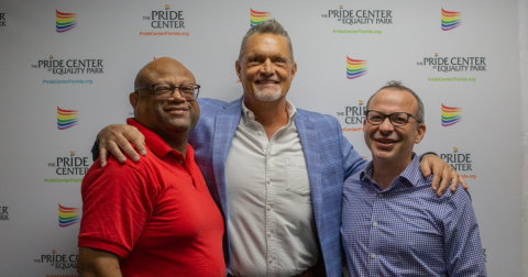 Three man in nice outfits pose in front of a banner with the logo of The Preide Center at Equality Park stamped on it