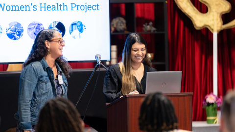 Two women smile at a podium while presenting from a computer. The screen in the background says "Women's Health Project"